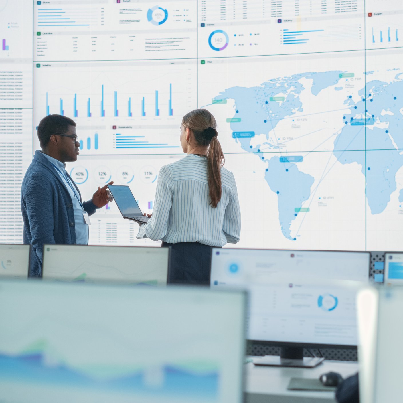 Black Male Analyst Talks To Caucasian Female Manager In Front Of Big Digital Screen With World Map In Monitoring Office. Diverse Employees Working Behind Desktop Computers In Logistics Company.