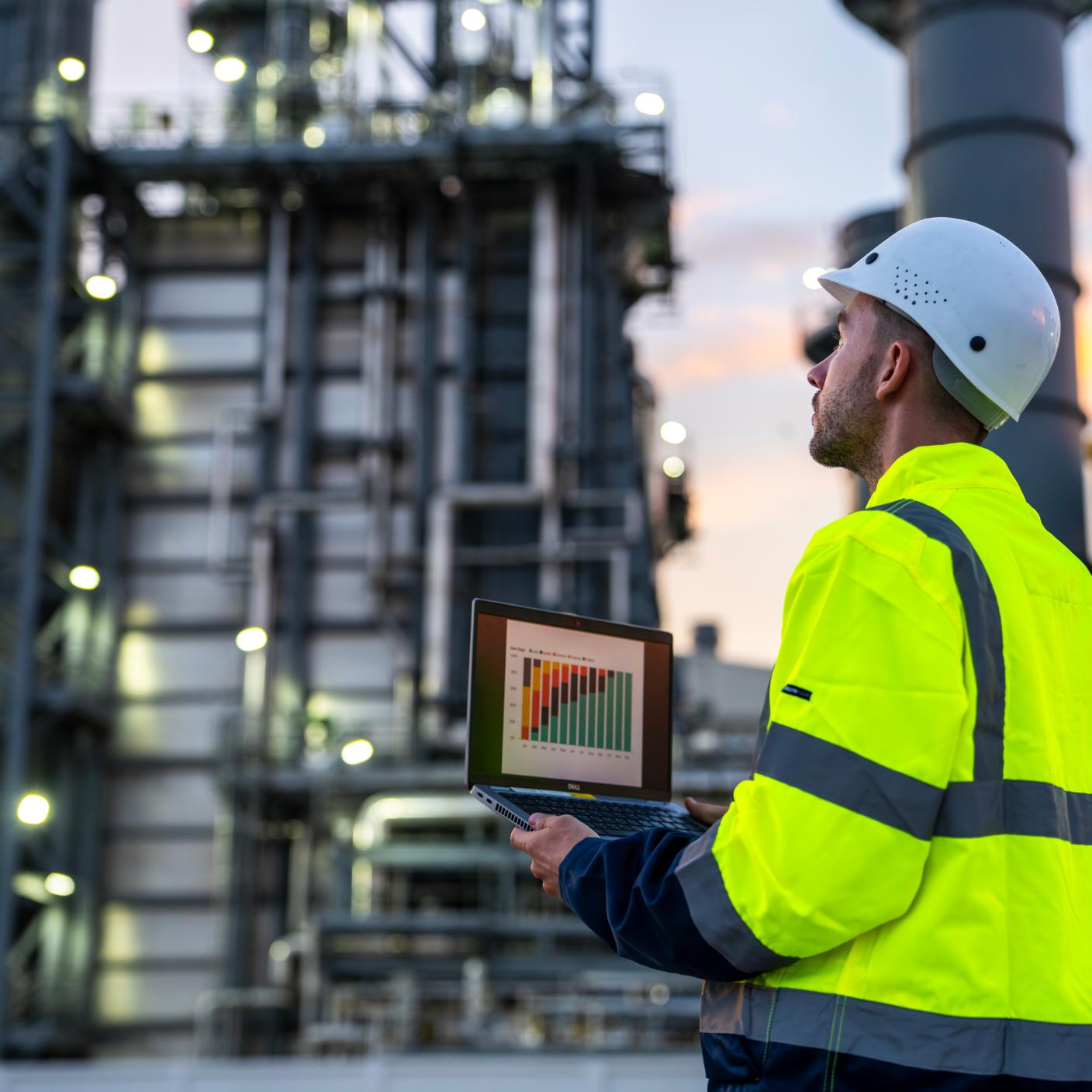 An industrial engineer using a laptop to monitor operational data at a facility. The scene portrays technology integration in modern engineering and manufacturing in a complex setting during the sunset.
