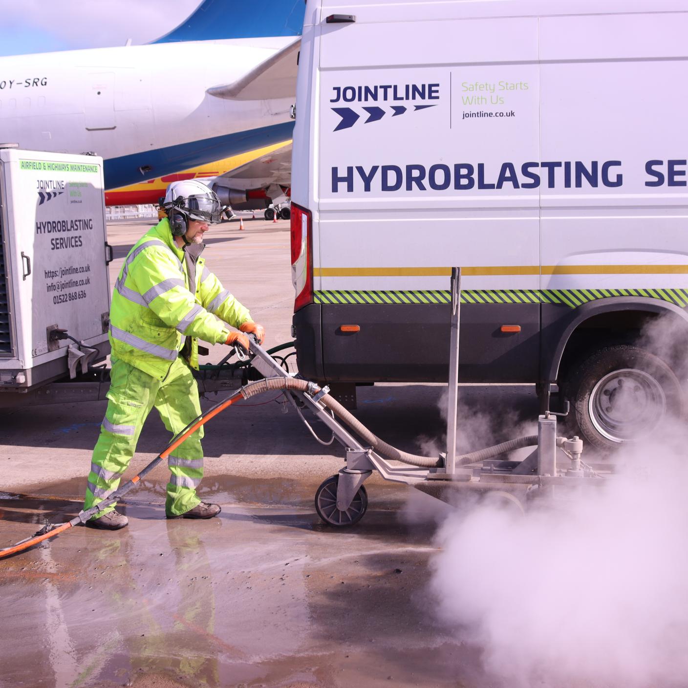 A worker in high‑visibility clothing operates a hydroblasting machine on an airport surface.
