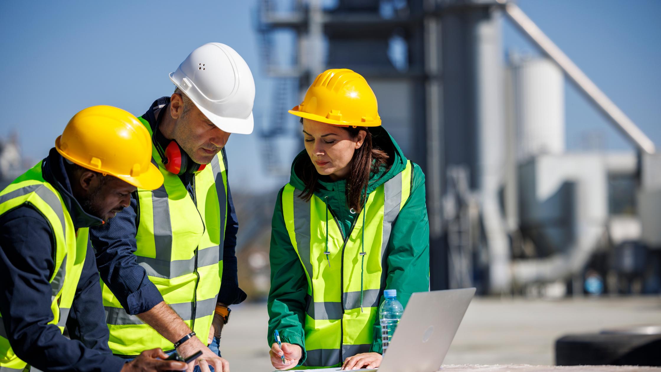 Three engineers in safety attire discussing at an industrial facility writing on paper next to a laptop.