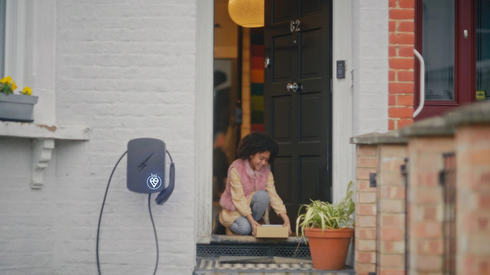 A young girl picks up a package at a front door, next to a wall-mounted home EV charging station with a BSI certification logo.